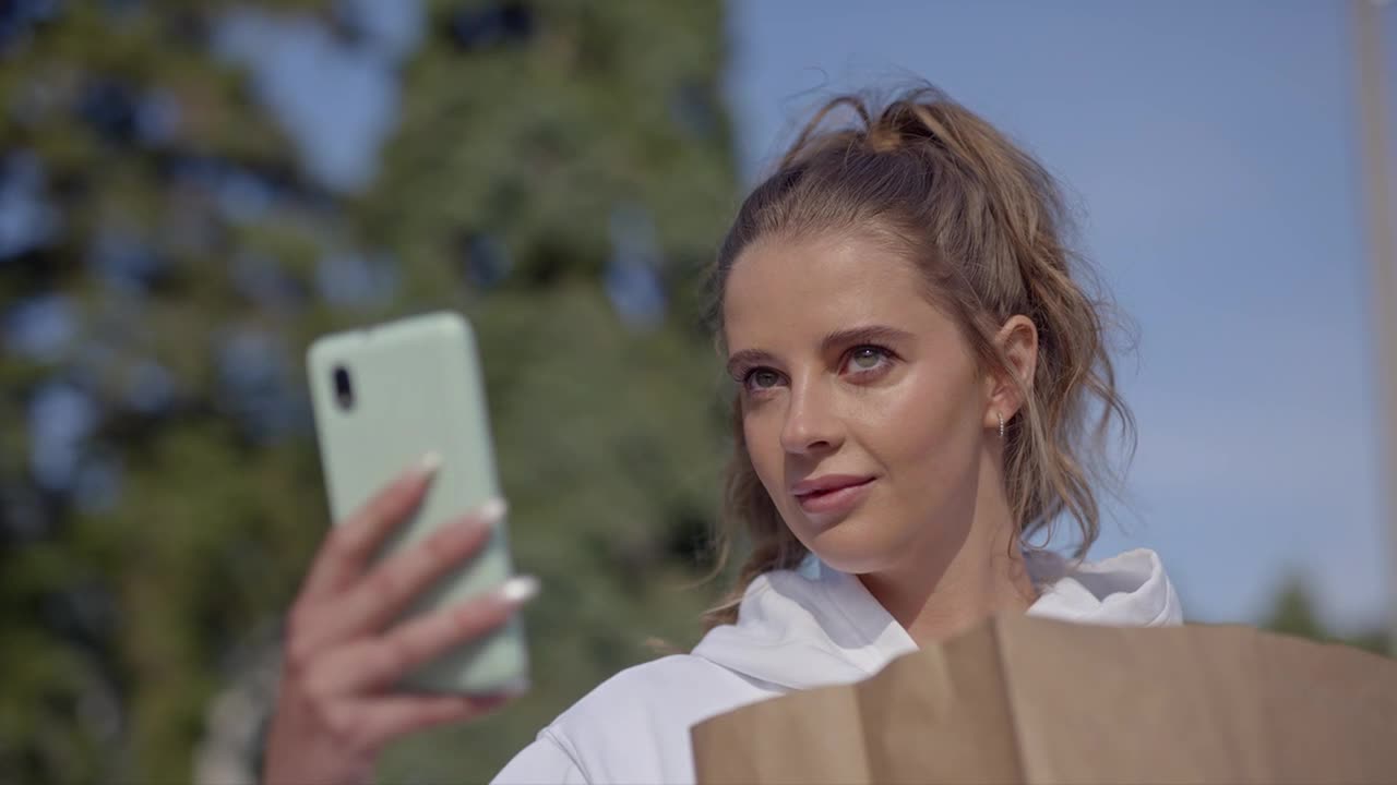 She's holding a phone up, angled to take a picture, and looking at the screen. A brown paper bag rests in her other arm, and she's outside in the sunlight.
