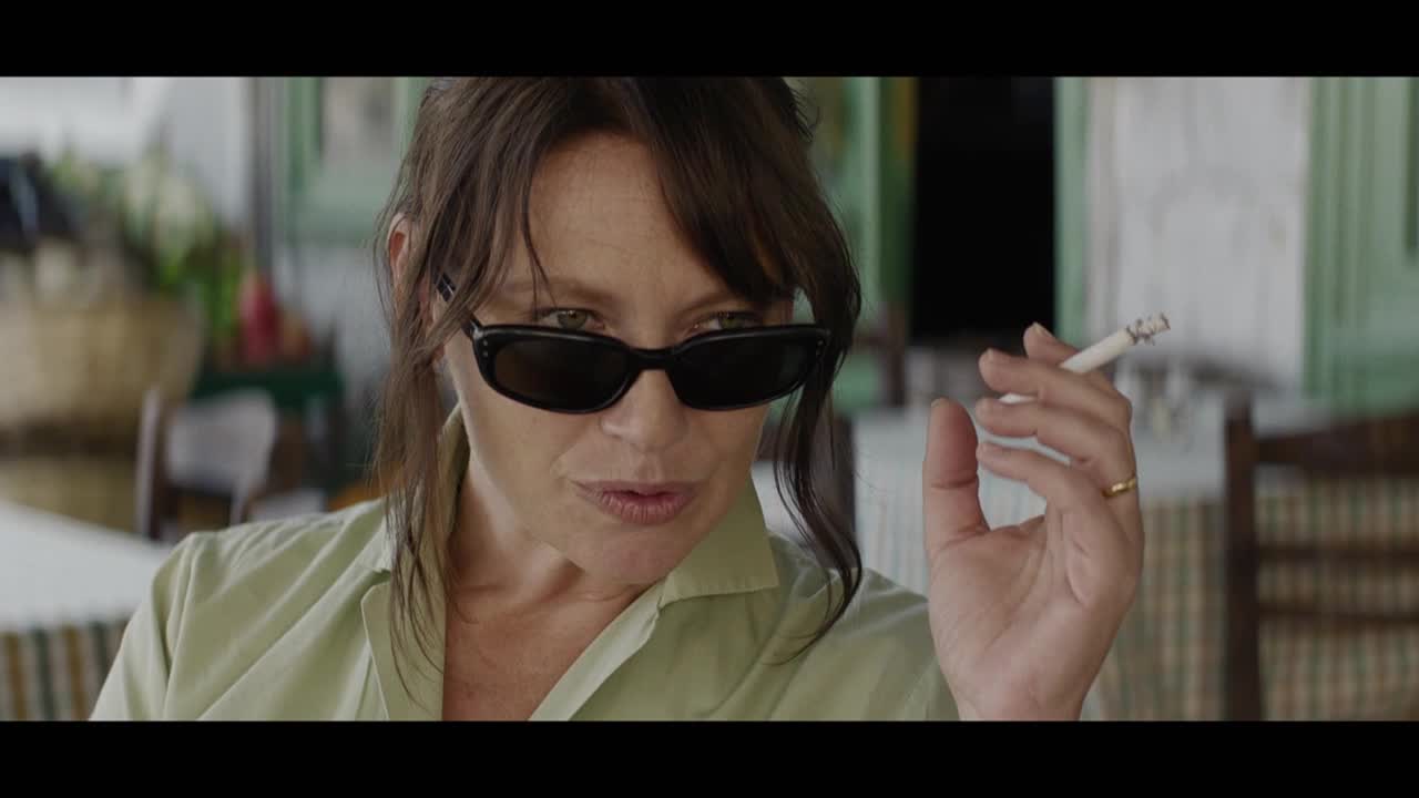 She's wearing sunglasses, squinting slightly as she holds a lit cigarette between her fingers. The smoke curls upwards, disappearing into the air.

