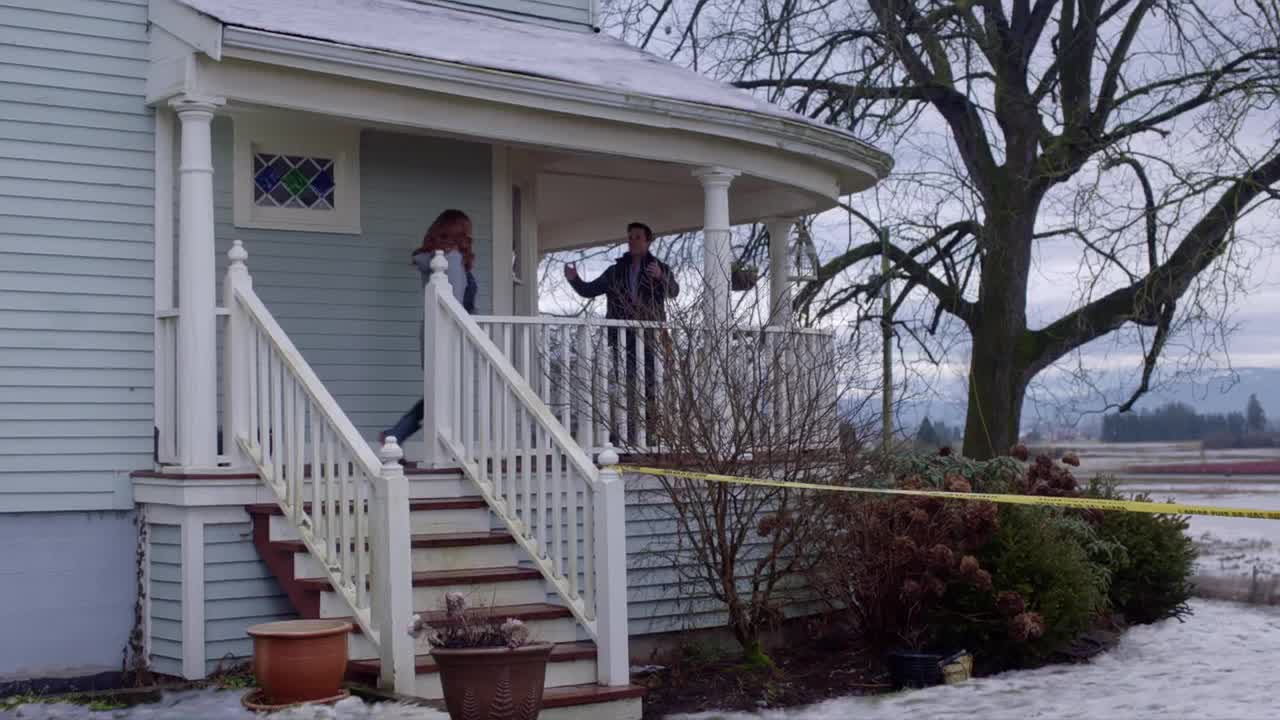 A woman descends the porch stairs, while a man gestures from the railing. Yellow tape marks the perimeter of the house, suggesting something has happened.
