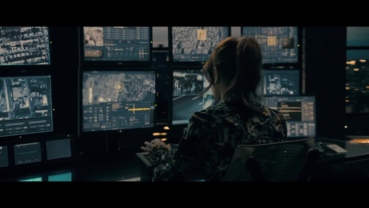 A woman with her hair pulled back sits in front of a wall of monitors, her fingers moving across a keyboard. The screens display maps and data, illuminated by the glow of the city outside the window.
