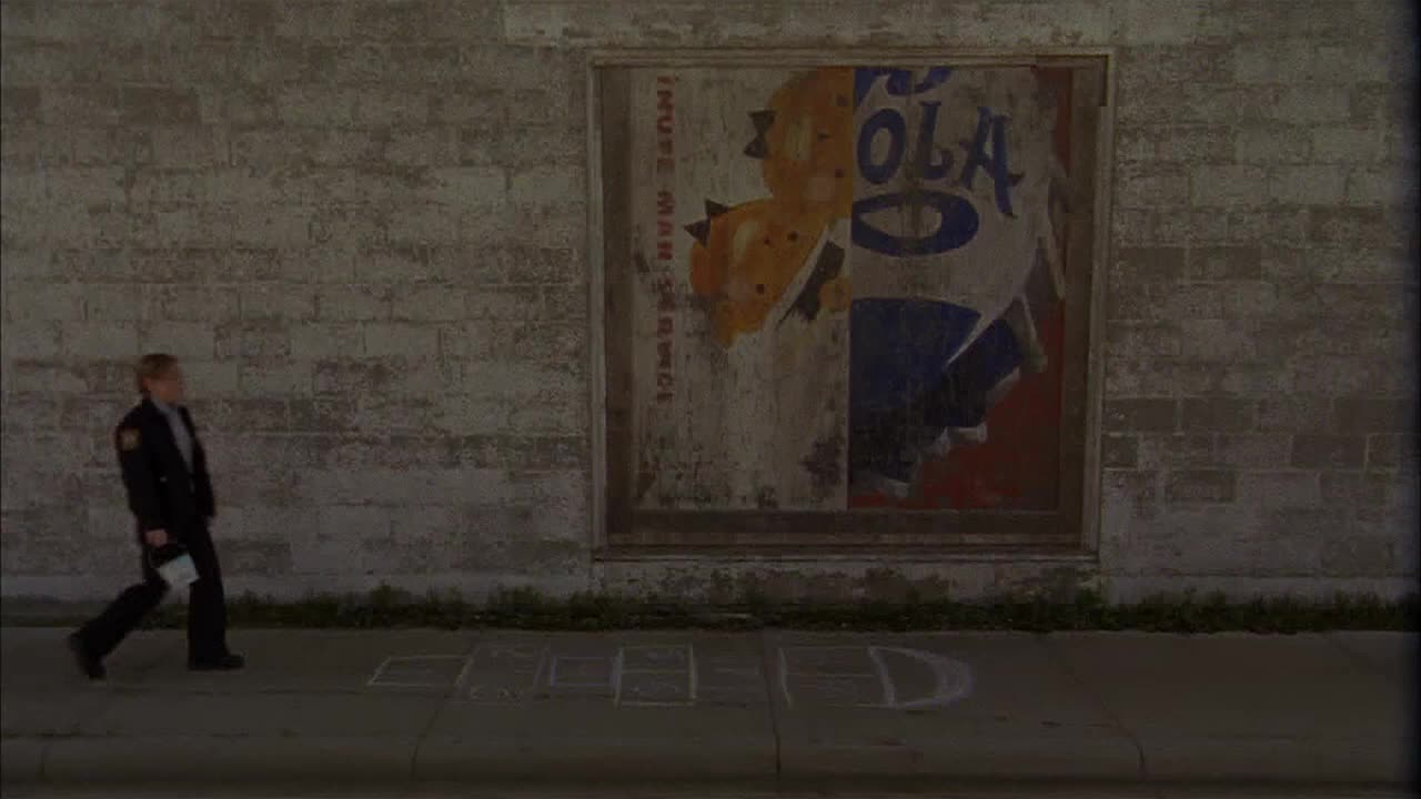 A man in a dark uniform walks past a faded, vintage cola advertisement on a brick wall. The asphalt sidewalk has a hopscotch game drawn on it.
