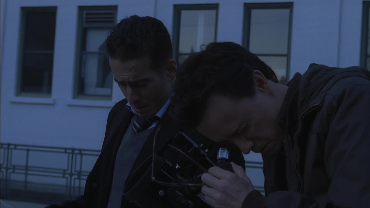 Two men huddle close, their faces illuminated by a dim light. One examines a small, black device, while the other looks on with a concerned expression.
