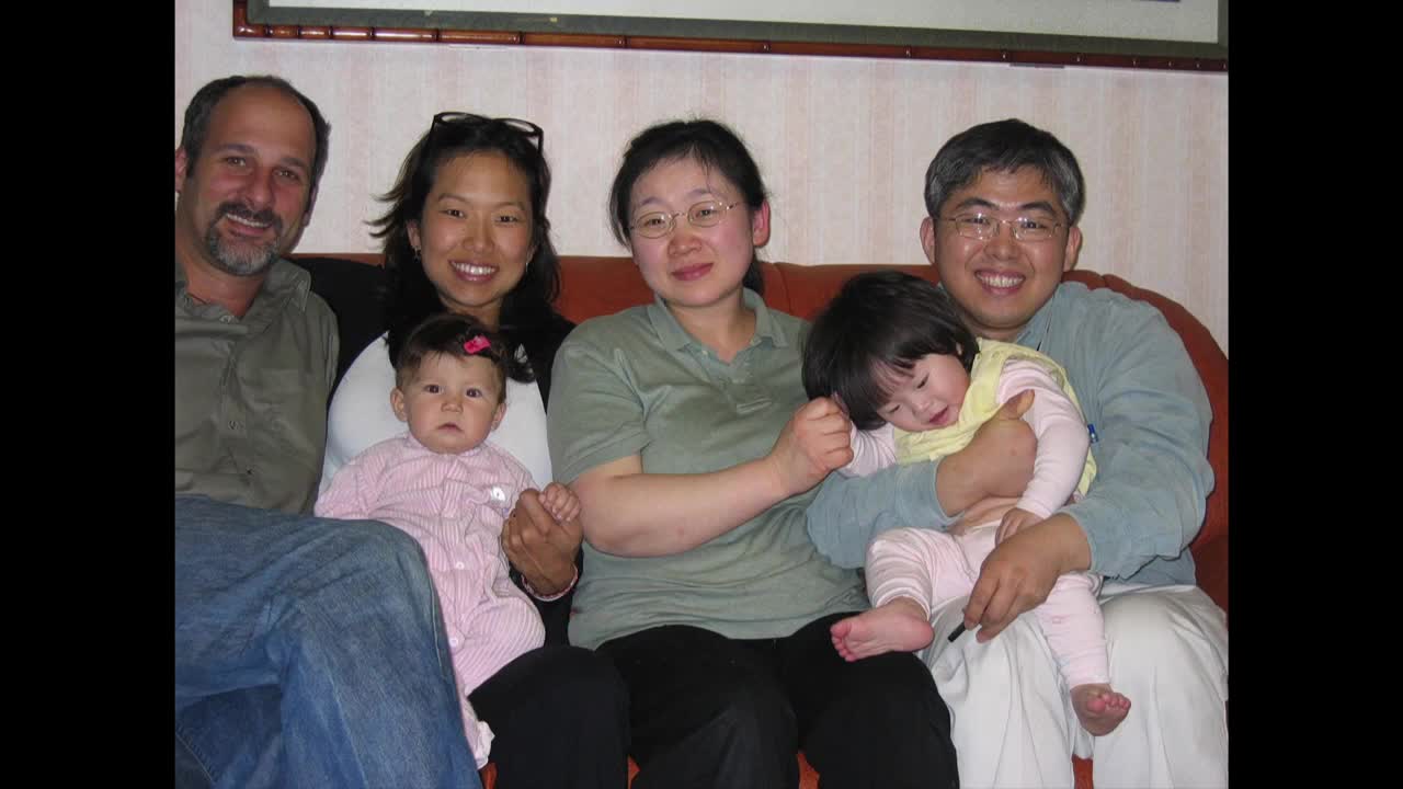 A group of six people are seated on a red couch, smiling at the camera. Two babies are held in their parents' arms, one looking directly at me, the other leaning against their dad.
