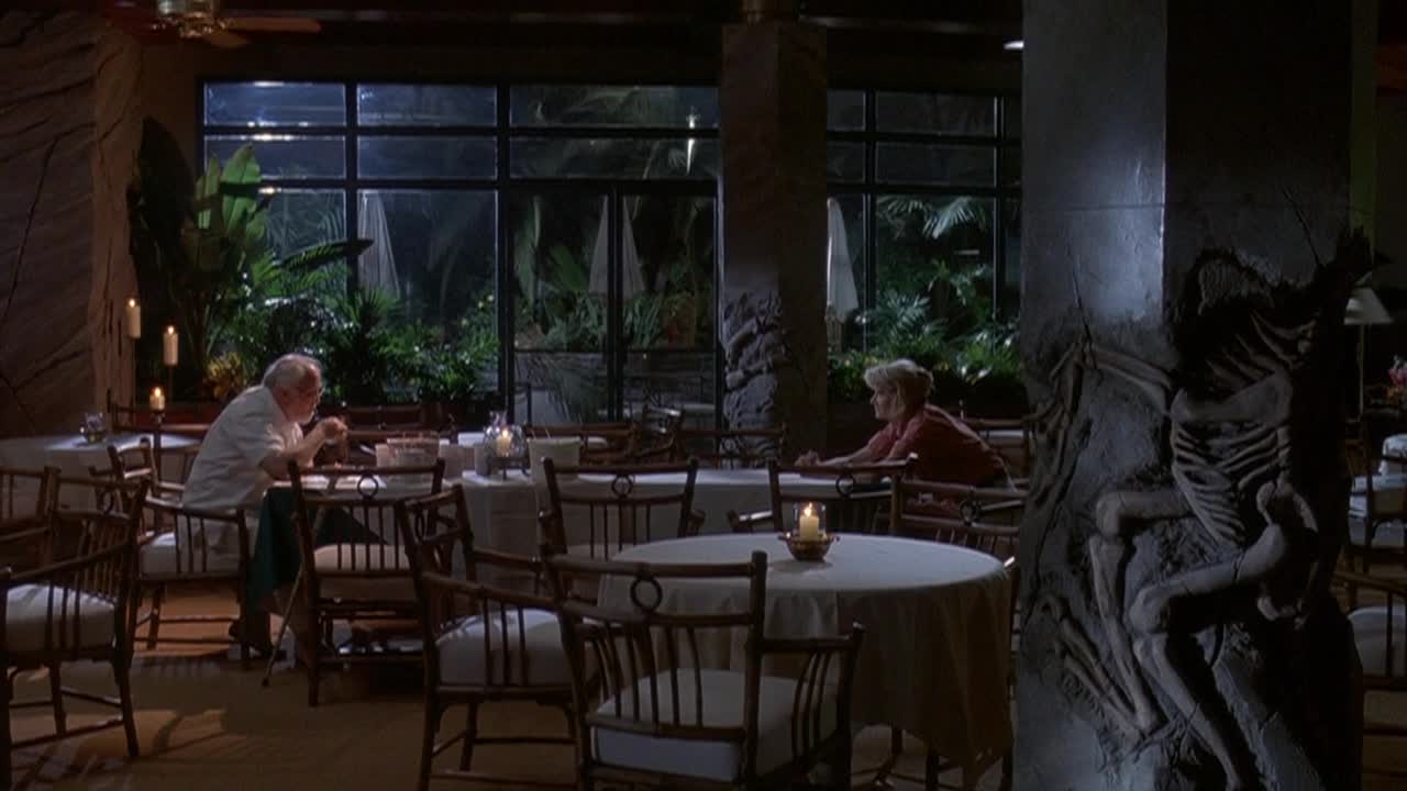 An older man in a white shirt sits hunched over a table, while a woman in a red top sits at a separate table, looking away. The restaurant, with its round tables and wicker chairs, is dimly lit by candles.
