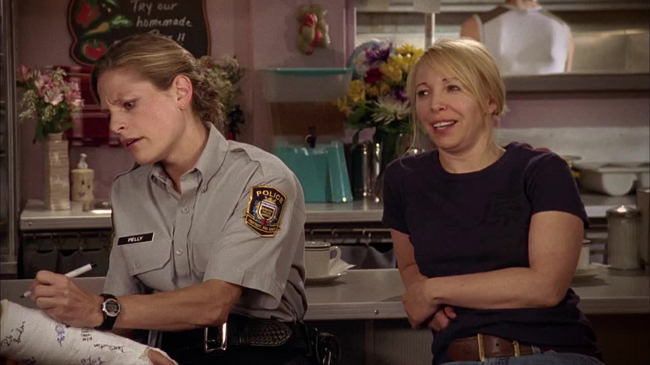 The police officer, Pelly, is writing on a cast, her brow furrowed in concentration. Across from her, a woman in a dark t-shirt watches, a small smile playing on her lips.
