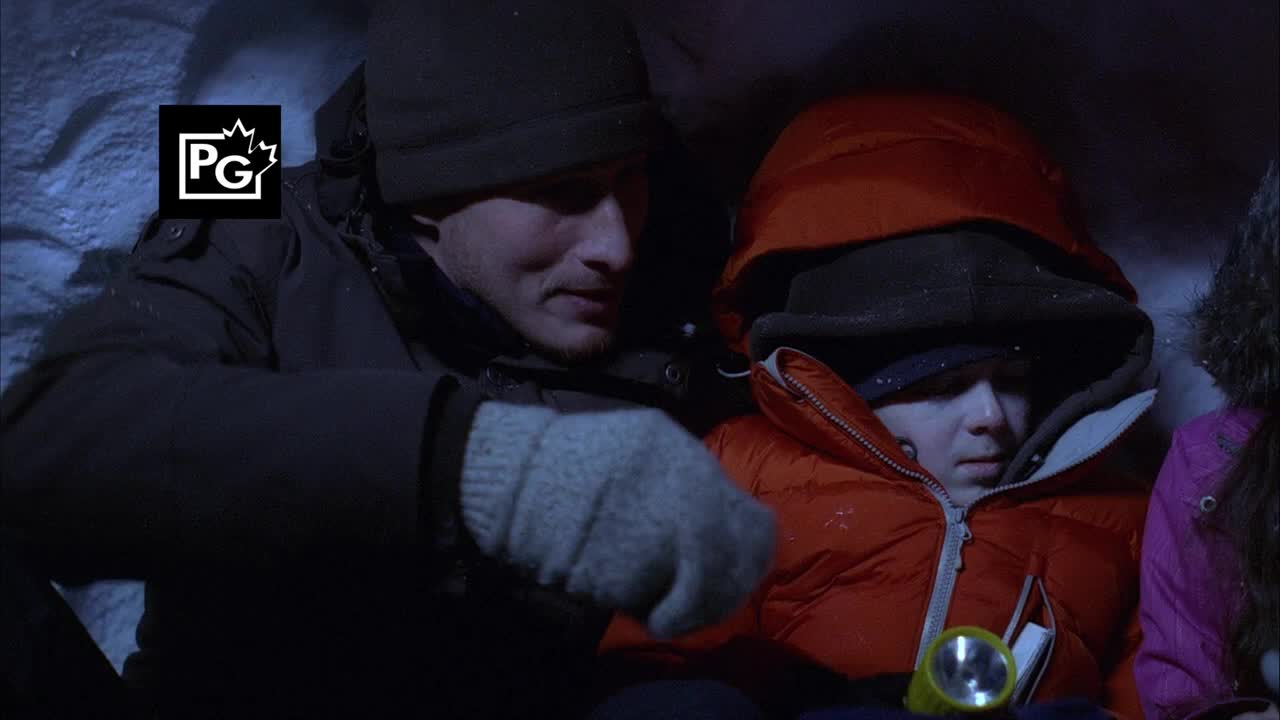 A man in a black hat and gloves leans toward a child bundled in an orange parka. A small flashlight rests on the child's lap, illuminating their faces in the dim space.
