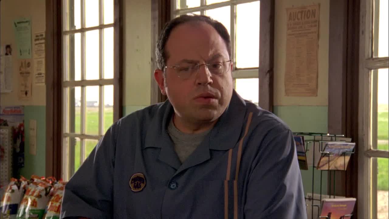 The man in the blue uniform looks concerned, his gaze fixed on something off-screen. He's standing in a small, old-fashioned shop, with windows overlooking a green field.
