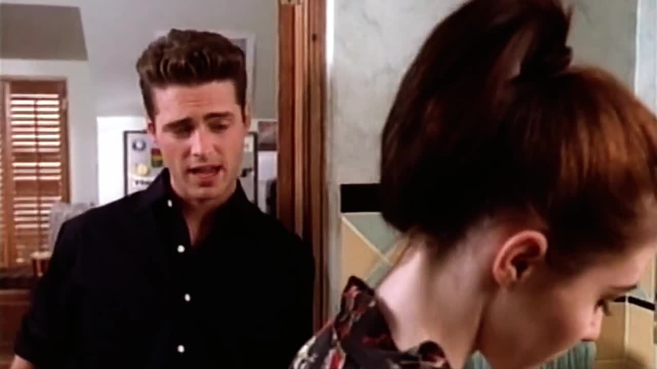 A man in a black shirt gestures with his hands as he speaks, his expression earnest. A woman with her hair pulled back is turned away, her face partially obscured.
