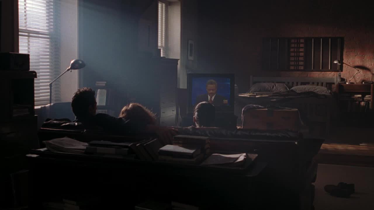 Two figures are slumped on a long, dark couch, their faces turned towards the TV. A news anchor on Crave 2 is speaking, the only source of light in the dimly lit room.

