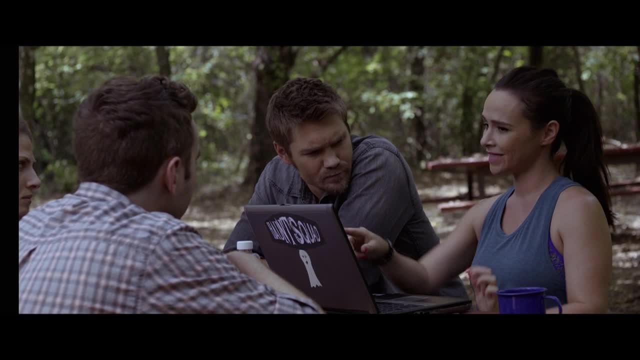 A group sits around a laptop with a "Haunt Squad" sticker, pointing and talking. The woman in the blue tank top gestures with her hand while the man in the middle leans in, focused.
