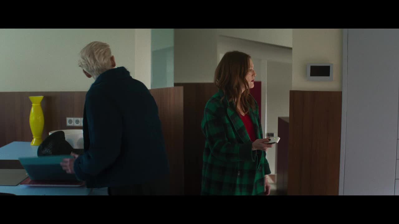 A man with white hair is hunched over a table, moving something into a container. Across the room, a woman in a green plaid coat holds a remote, looking on with concern.
