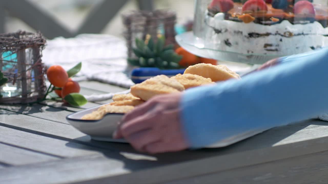 A hand in a blue sleeve is setting down a plate of golden, square-shaped cookies on a weathered wooden table.  Behind the plate, a tiered cake covered in berries sits under a glass dome.
