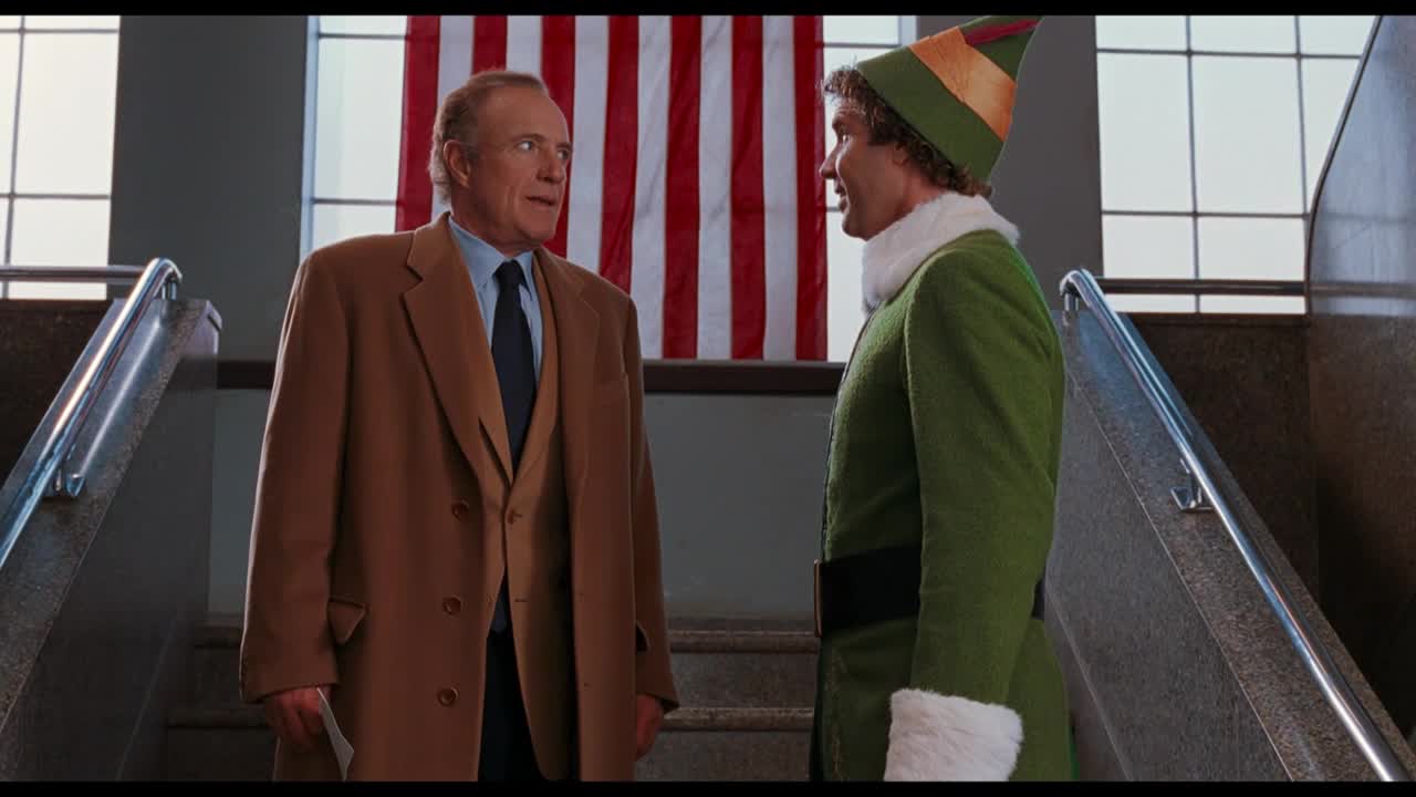 A man in a suit stands facing an elf in a green costume, both on a staircase. The elf is animatedly speaking to the man, who appears to be listening with a slight smile.
