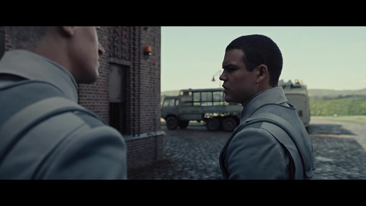 Two men in matching gray uniforms stand facing each other near a brick building, their expressions serious. Beyond them, a military truck sits parked on a cobblestone surface, with fields stretching out in the distance.
