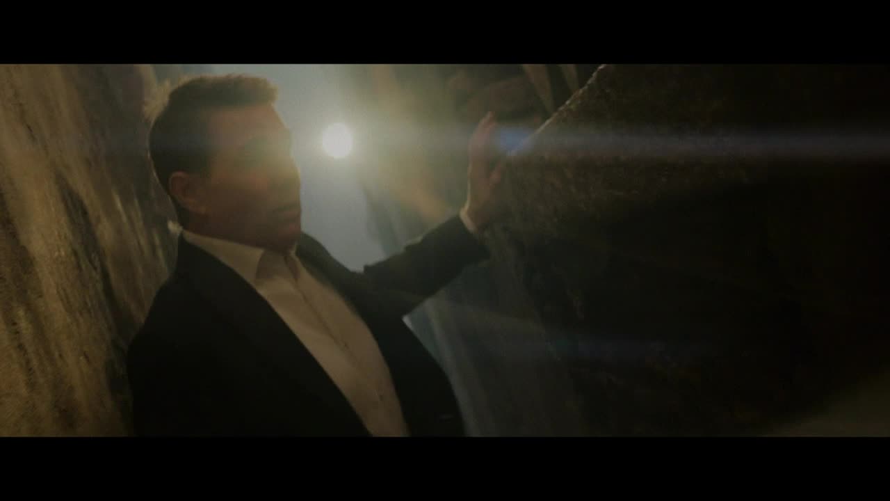 A man in a suit reaches out, his hand pressed against a rough, dark stone surface. A bright light shines from behind him, obscuring his face.
