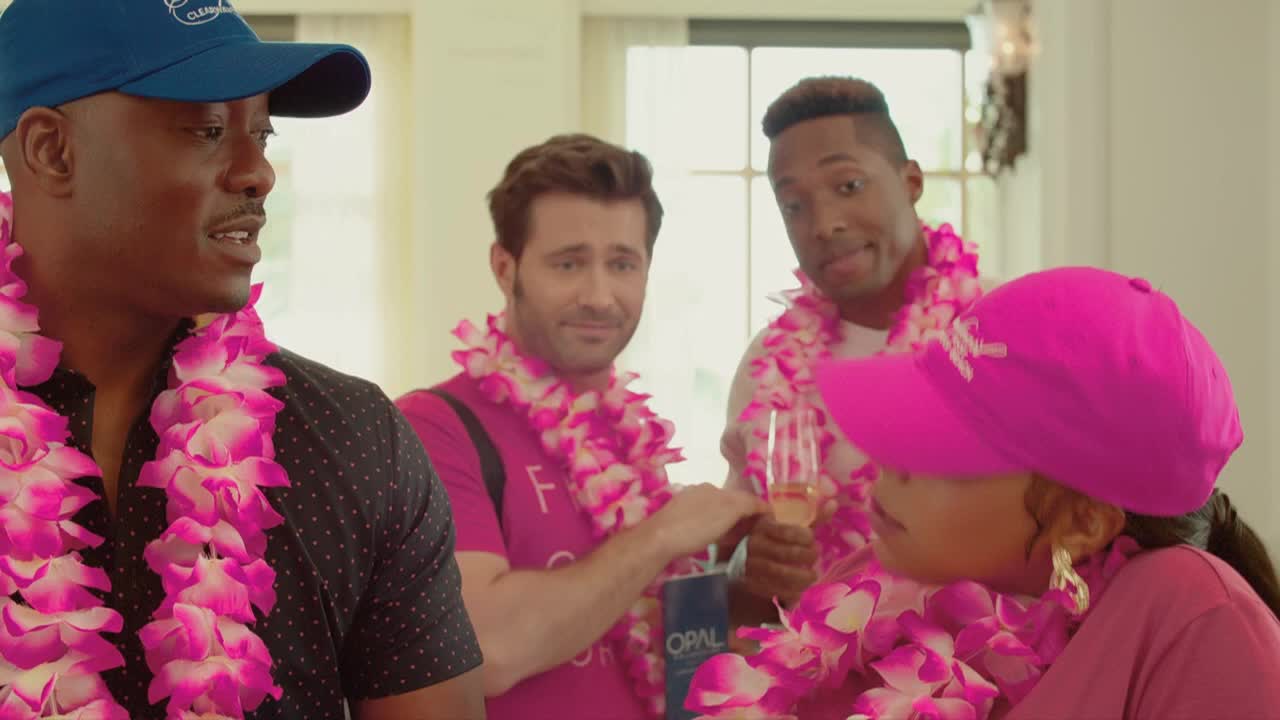 Four people, all wearing leis, are gathered in a room, with two holding glasses of champagne. The man in the blue cap is speaking, while the others appear to be listening.
