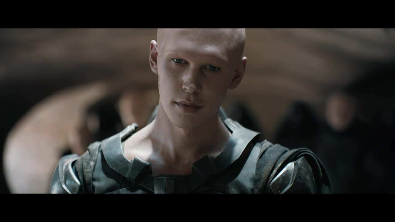 The man with the shaved head is looking directly at me, his expression unreadable. He's wearing a dark, angular garment, and the lighting casts a shadow over the room.
