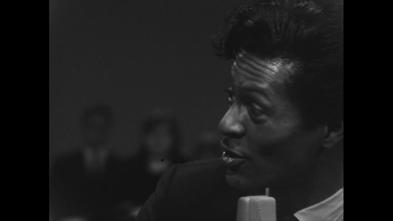 The man is speaking into a microphone, his face lit up by the studio lights. Behind him, a few indistinct figures sit in the shadows.
