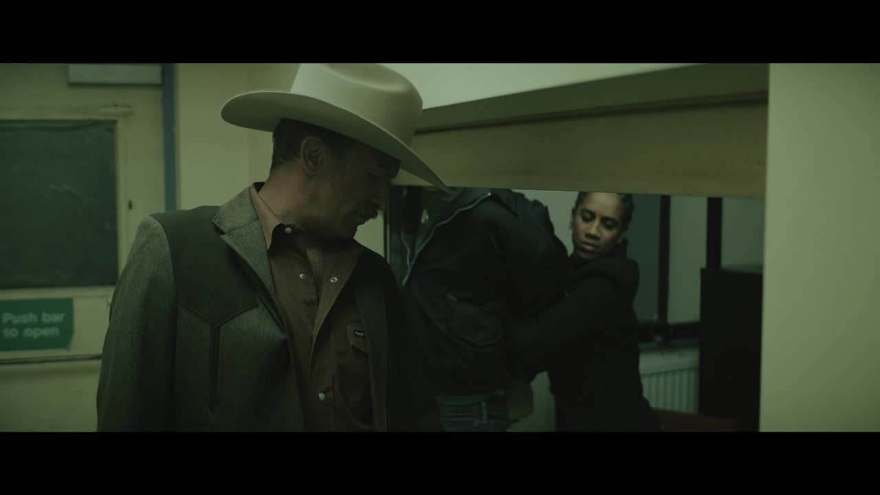 The man in the cowboy hat looks down as two people struggle out of frame. A woman in a black jacket is holding someone, their back to the camera.

