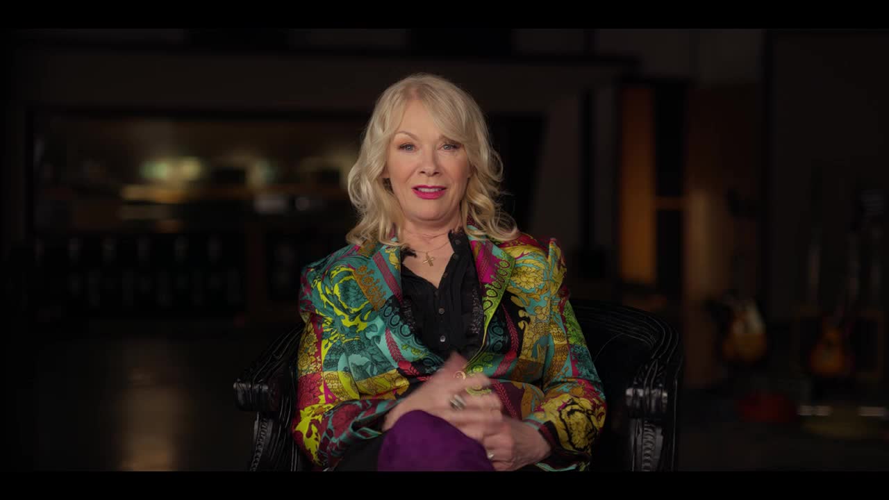 A woman with blonde hair and a colorful jacket sits in a chair, her hands clasped in her lap. She's looking directly at the camera, probably for an interview on Crave 2 from Canada.
