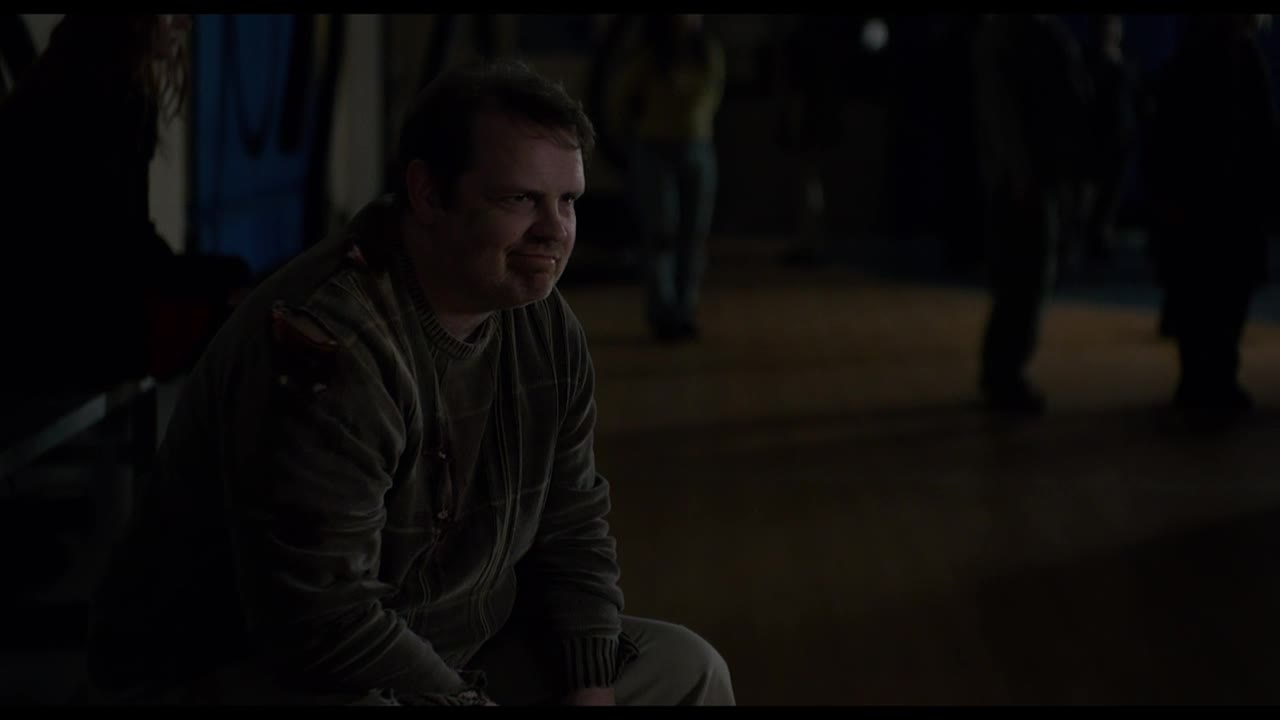 A man in a worn-out zip-up sweatshirt sits, looking off to the side with a slight smile. Behind him, figures are indistinct, and the lighting is dim.

