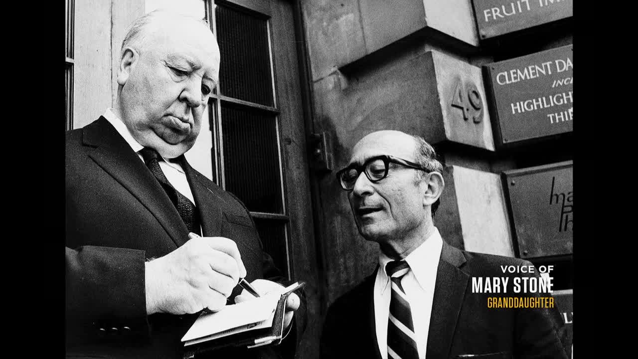 Hitchcock, pen in hand, is focused on his notepad. Beside him, a man with glasses listens intently, seemingly part of the conversation.
