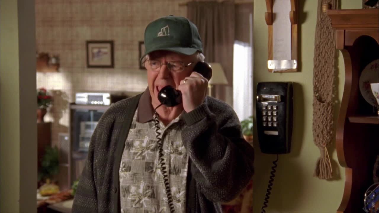 An older man in a green cap is holding a black phone receiver to his ear, his mouth slightly open. He's standing in a cozy, well-lit living room, likely in Canada, as seen on Crave 2.
