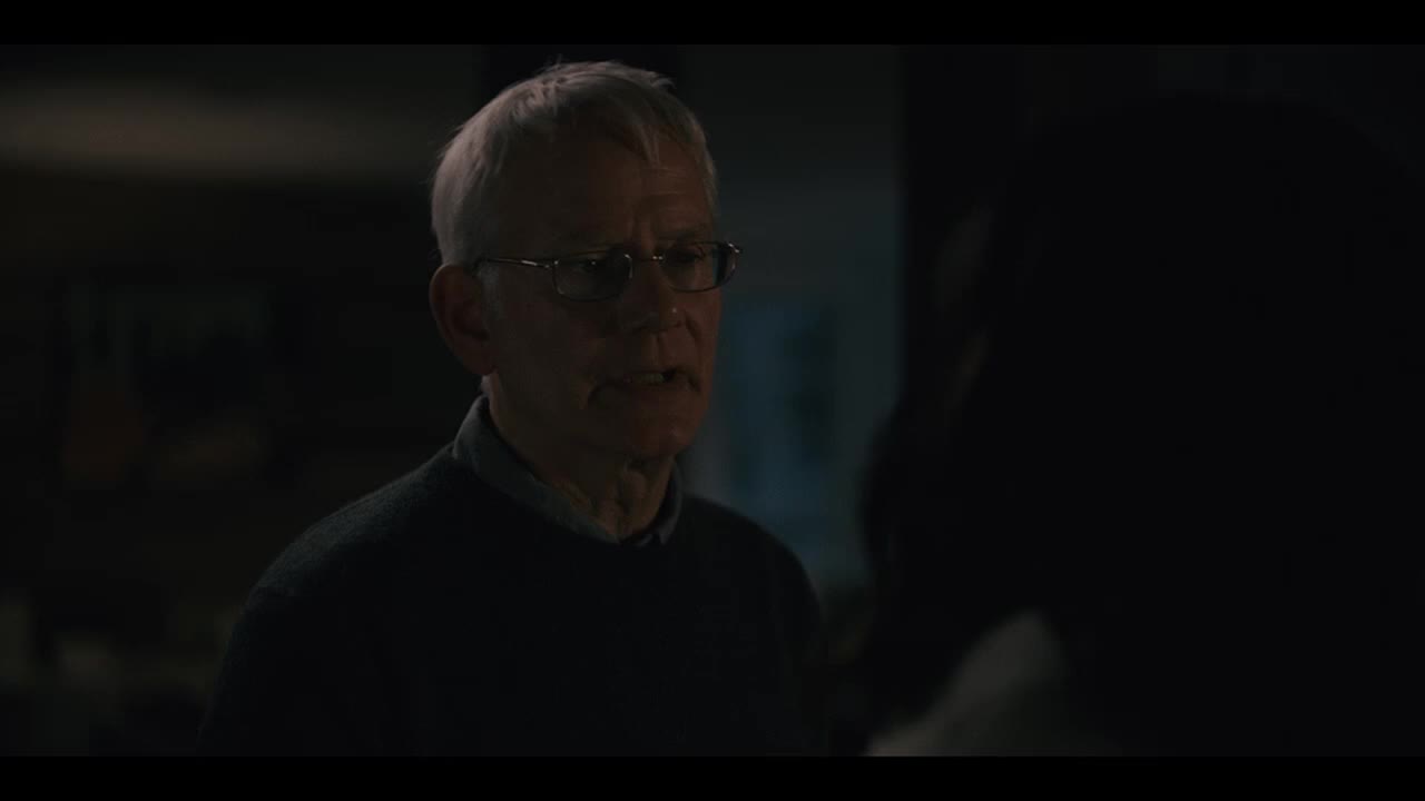 The older man, wearing glasses and a dark sweater, is speaking intently. His gaze is fixed on the person off-screen, who is mostly in shadow.
