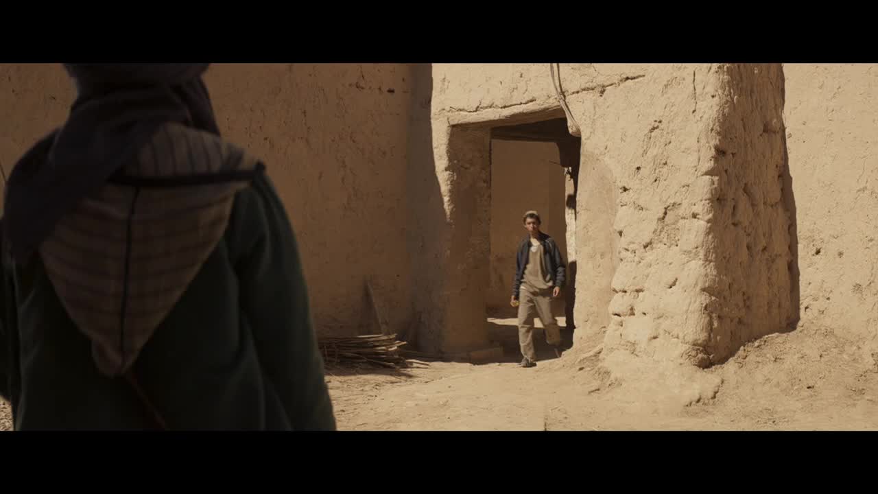A man in a jacket and khakis walks out of a doorway, heading toward the camera. Someone in a hooded cloak stands with their back to me, watching him.
