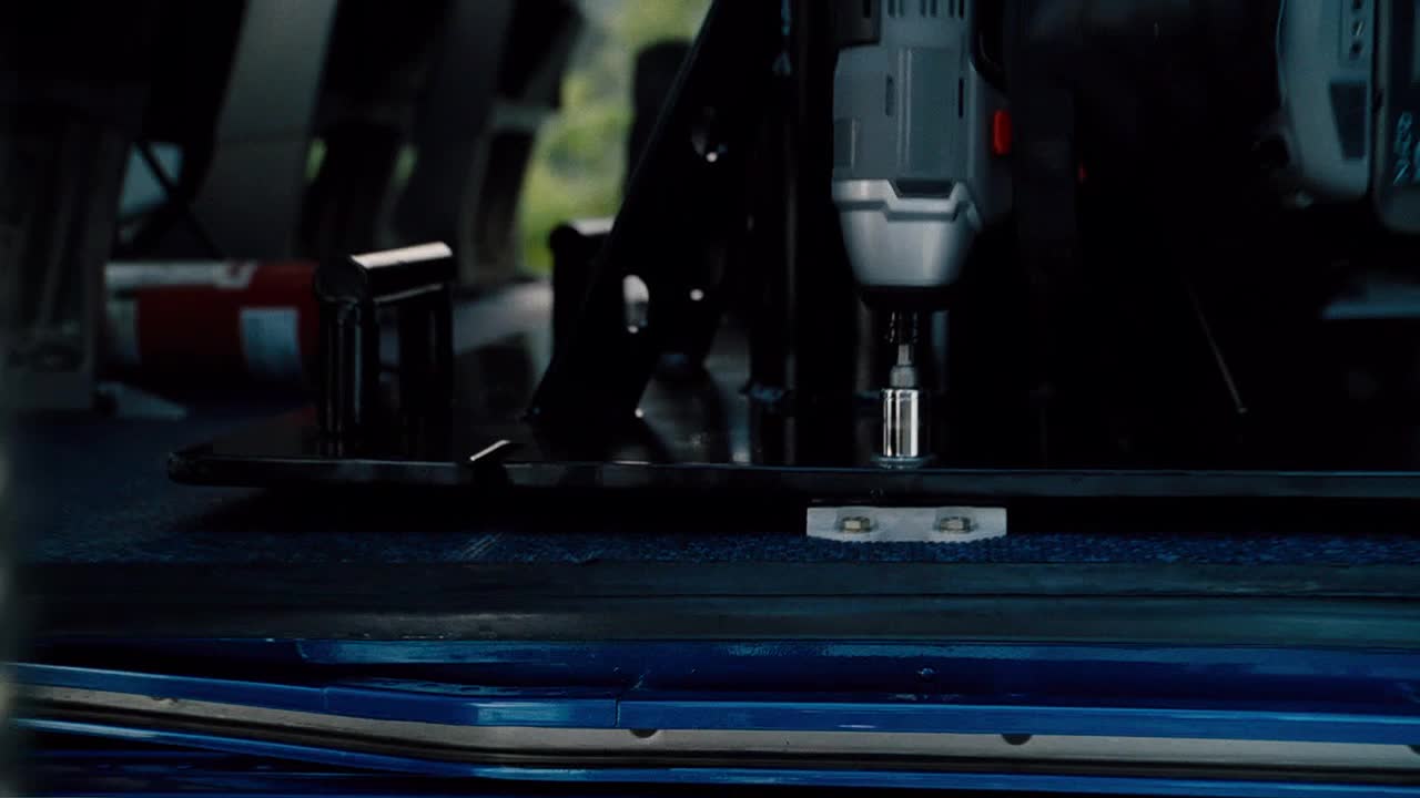 A power drill is held over a metal plate, its silver bit poised to screw something in. The tool is angled, and the work is happening inside a vehicle.
