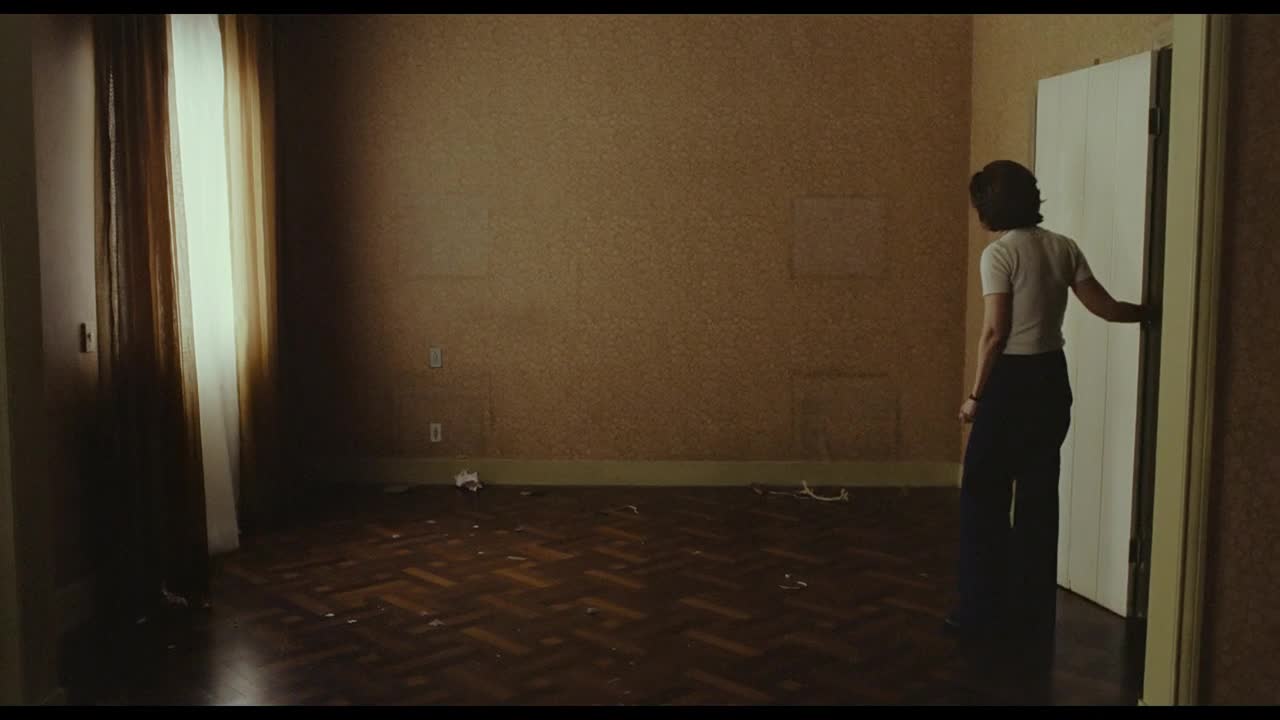 A woman in a white shirt and wide-legged pants stands in a sparsely furnished room, her hand resting on the doorframe. Light streams in from a window, illuminating the dusty floorboards and the peeling wallpaper.
