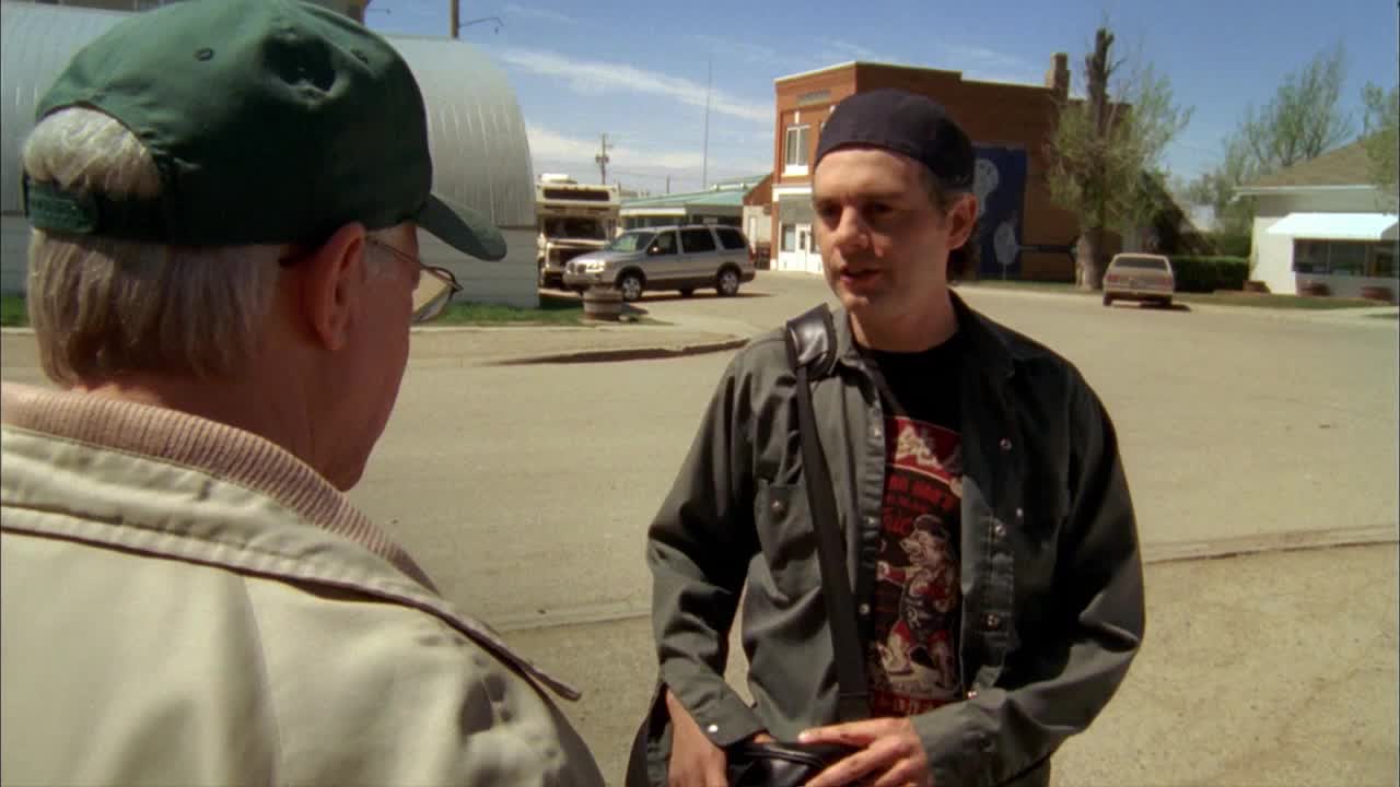 A man in a black cap gestures while talking to someone wearing a green baseball cap. The scene is set on a quiet street in a small Canadian town.
