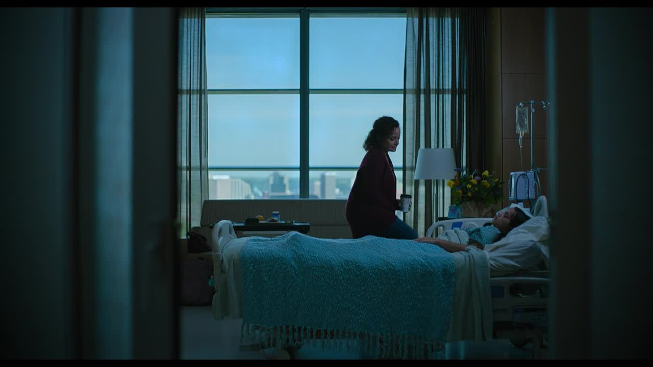 A woman sits on the edge of a hospital bed, looking down at the patient. The room is lit by a large window overlooking a cityscape, and a lamp casts a warm glow.
