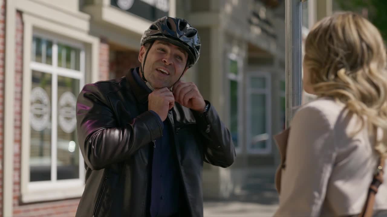 A man in a black leather jacket is adjusting his bike helmet, smiling at someone off-camera. His gaze is fixed on a woman with blonde hair, who seems to be waiting.
