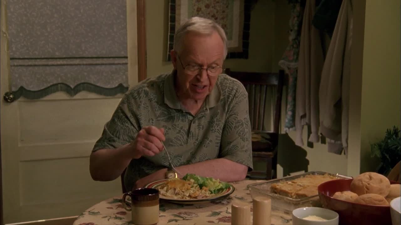 A man with glasses is using a fork to bring food to his mouth. He's at a table set for a meal, and there's a casserole dish and rolls nearby.
