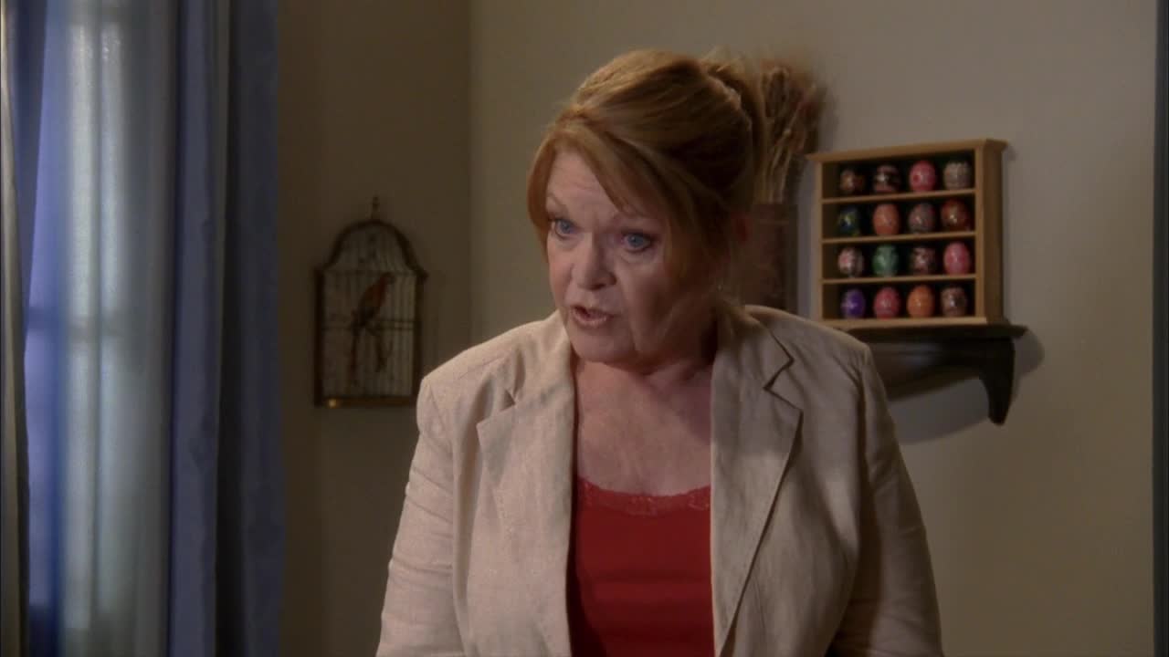 The woman in the blazer is mid-sentence, her expression serious. Behind her, a collection of decorative eggs sits on a shelf.
