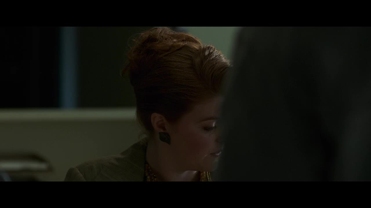 A woman with auburn hair, styled in a high updo, looks down and to her right, her expression unreadable. She wears a green jacket and a dark earring, the scene bathed in a dim, cool light.
