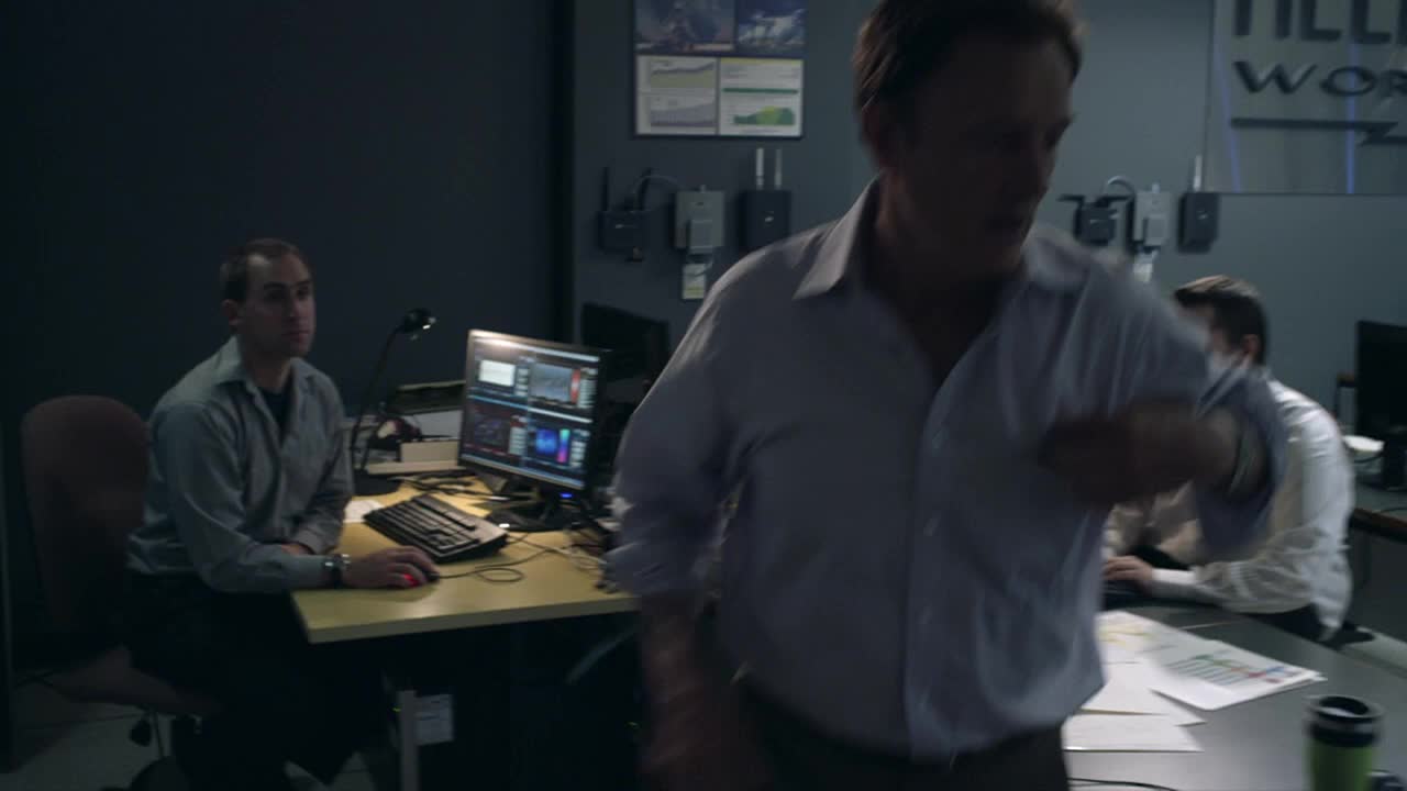 A man in a light blue shirt strides through the room, glancing at his watch. Two other men sit at desks, focused on computer screens and paperwork.
