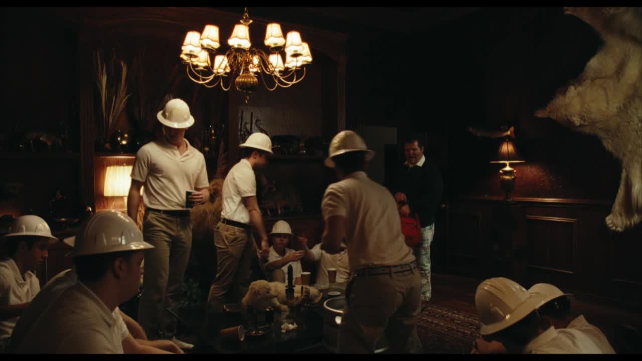 Men in white polo shirts and hard hats mill around a table, some holding drinks, in a dimly lit, wood-paneled room. A large bear skin hangs on the wall, and a chandelier casts a warm glow over the scene.
