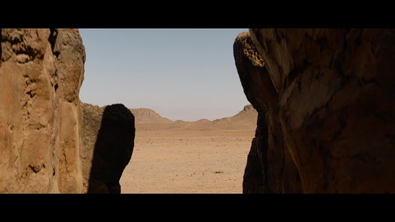 The desert stretches out, framed by two massive, sun-baked rocks. The distant hills are a hazy brown under a pale blue sky.
