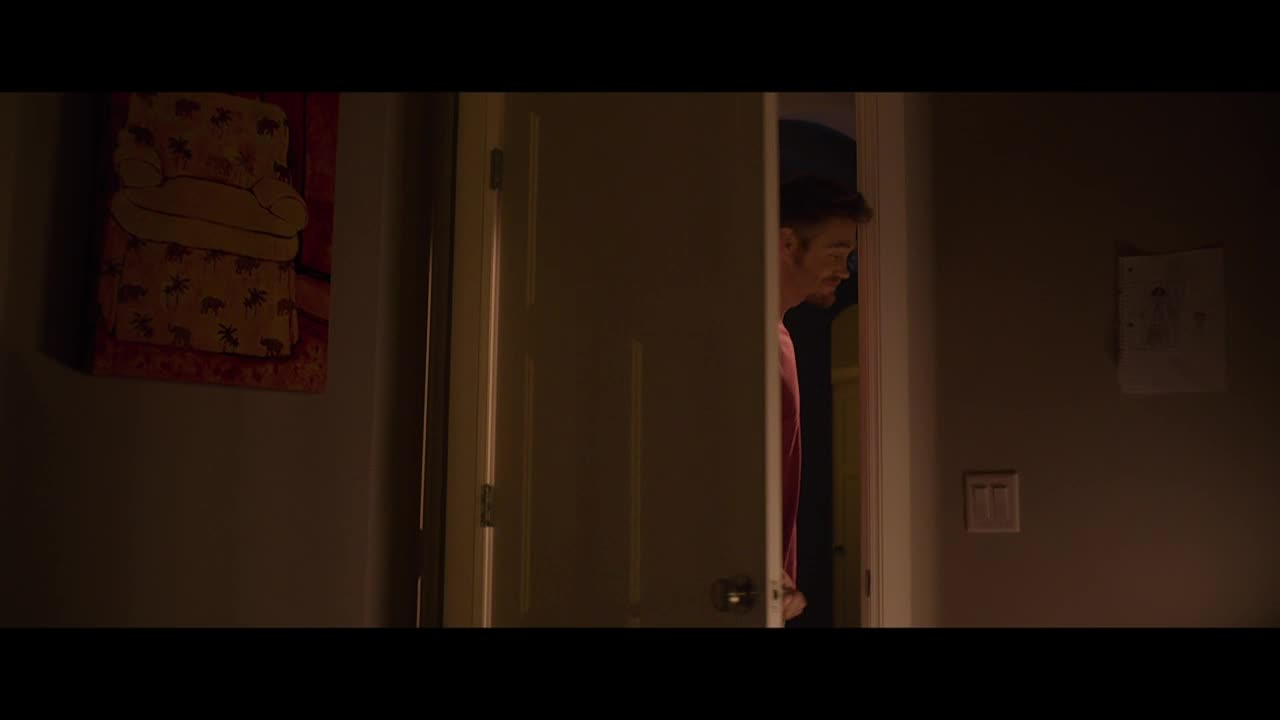 A man, wearing a red shirt, peers through a doorway, his face illuminated by an unseen light source. The room is dimly lit, with a child's drawing and a framed picture on the wall.
