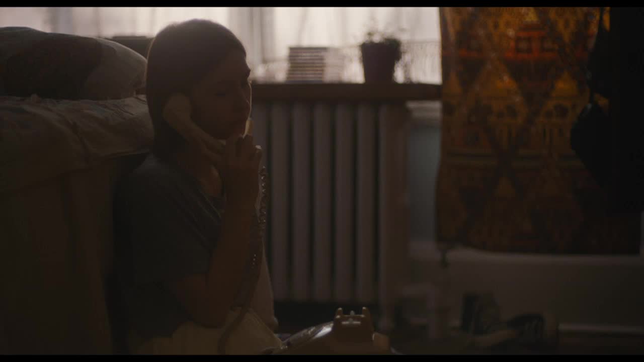 A young woman sits on the floor, the phone pressed to her ear. Sunlight streams through the window, illuminating the cord that snakes across the floor to the rotary dial phone.
