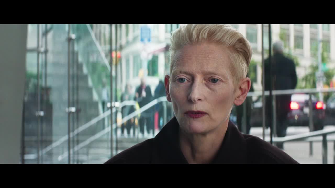 She's looking directly ahead, her gaze steady, with a city street reflected in the glass behind her. The light catches her short, blonde hair, and her dark lipstick stands out against her pale skin.
