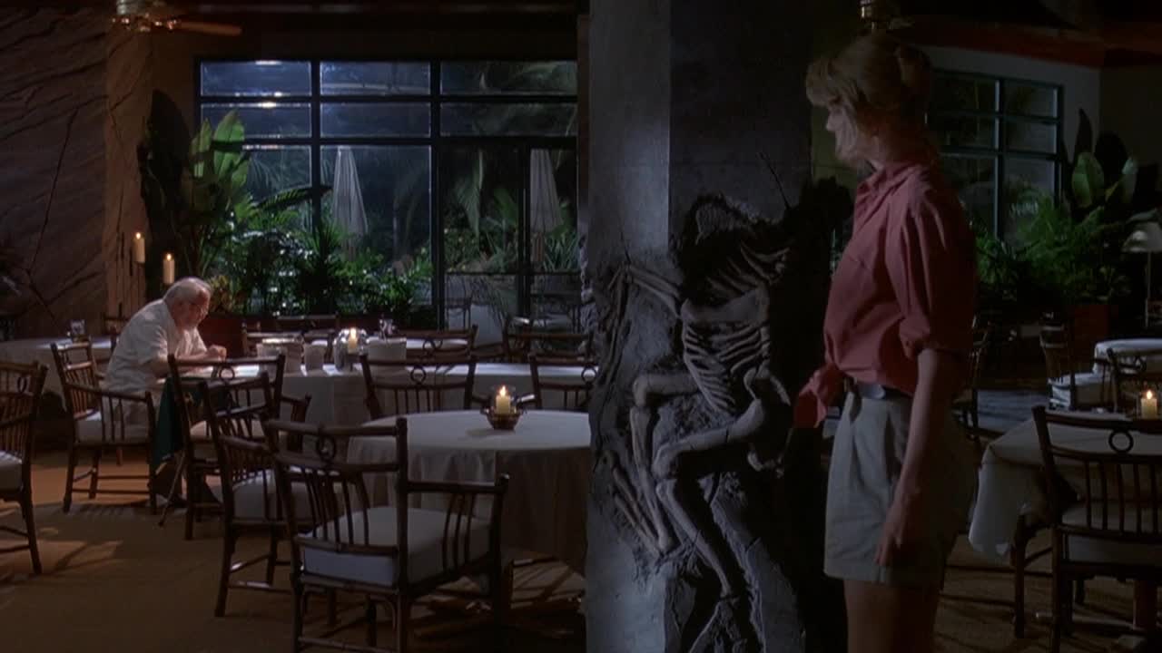 A woman in a red shirt and khaki shorts stands near a decorative pillar, her gaze fixed on something out of view. Across the room, a man sits alone at a table, illuminated by a small candle, as if he's waiting for a meal.
