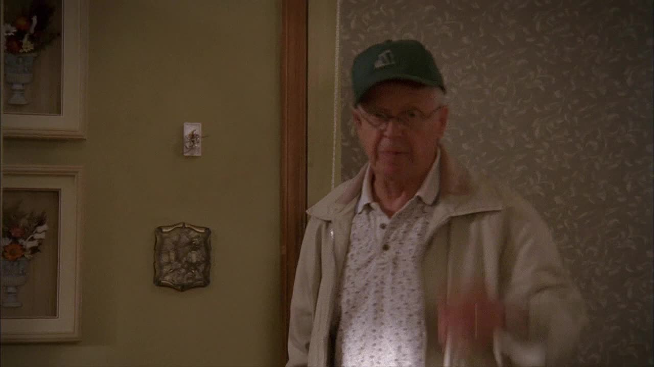 A man in a green cap and glasses gestures with his right hand, standing in a doorway. He's wearing a light jacket over a patterned shirt, and the scene feels like a quiet moment from a Canadian TV show on Crave 2.
