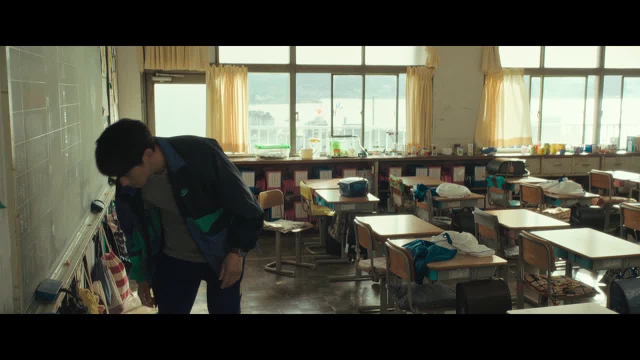 A man in a jacket is hunched over, examining something on a whiteboard in a classroom. Sunlight streams in through the large windows, illuminating the empty desks.
