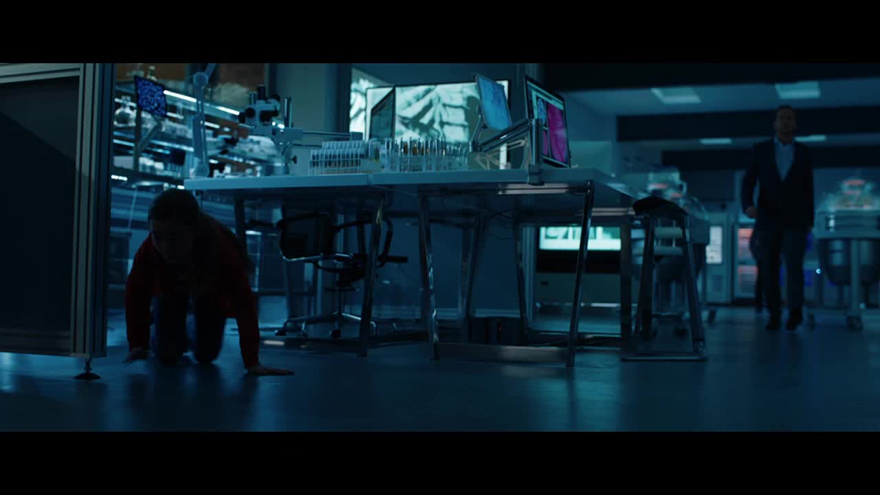 A person in a red shirt crawls low to the ground under a lab table. A man in a suit stands further back, watching.
