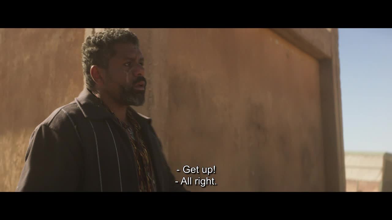 The man in the dark jacket, with a scar on his face, looks up and speaks, his words appearing as subtitles. The scene takes place outdoors, with a weathered brown wall behind him.
