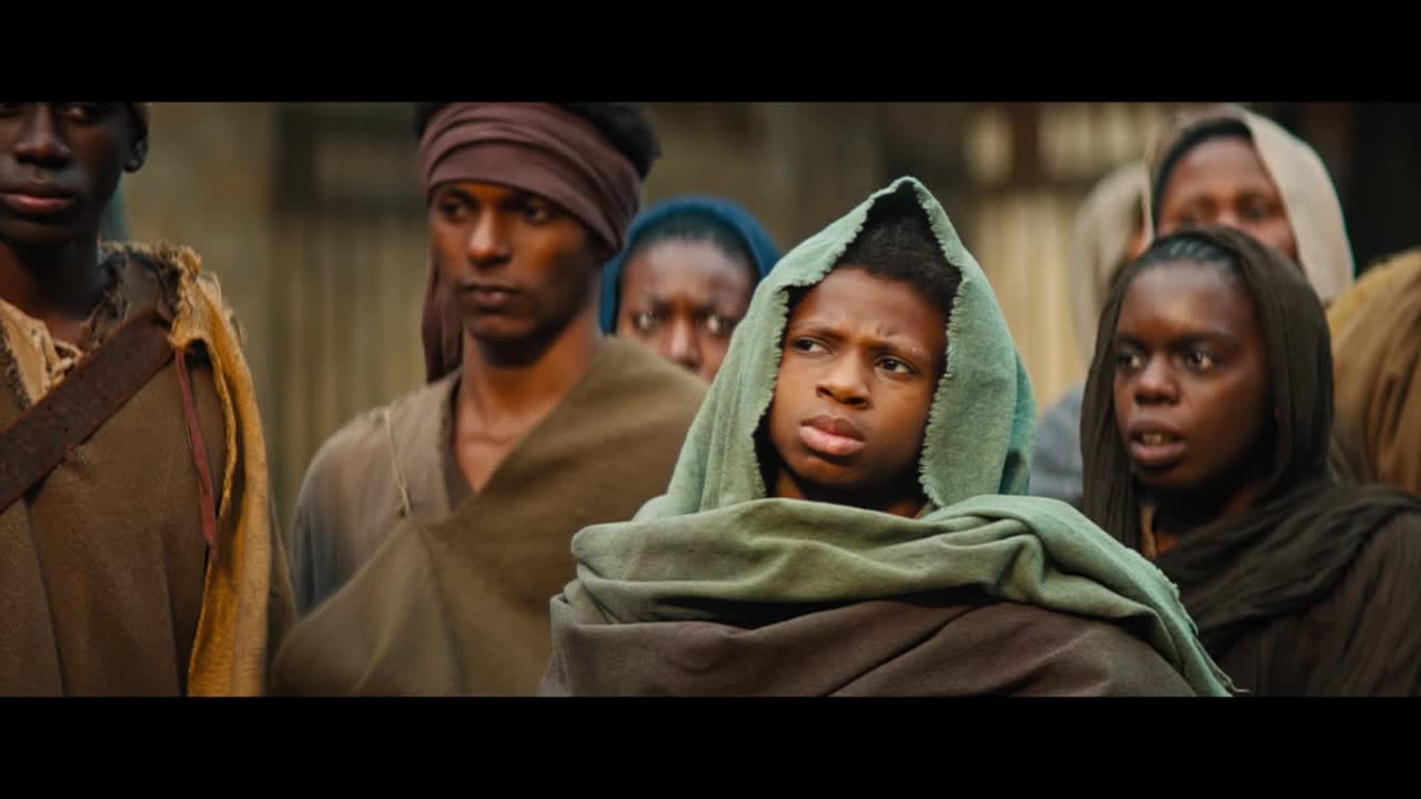 A group of people, wrapped in simple cloth, stand close together, their faces etched with concern. The young person in front, draped in green, looks directly forward with a furrowed brow.

