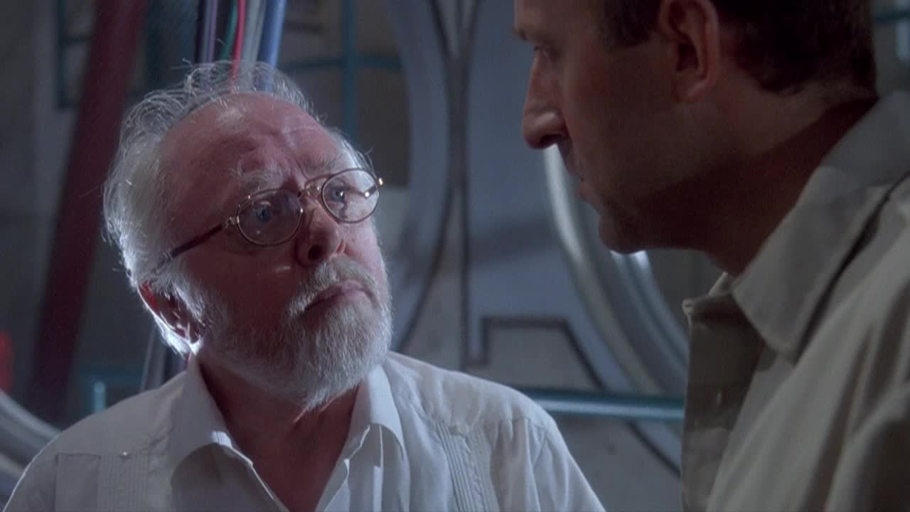 The older man, with a white beard and glasses, looks up, his eyes wide. He's listening intently to the other man, who's speaking just out of frame.
